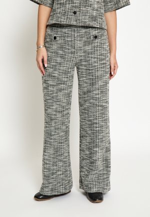 Kvinde iført sort-hvide teksturerede viddebukser og matchende kort jakke, stående med hænderne afslappet ned langs siderne, sorte sko synlige.