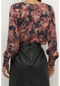 Blouse à manches longues bleu marine avec un motif floral aux tons roses et orange, dotée de poignets froncés et d'un détail dos en forme d'œillet. Portée avec une jupe en cuir noire.