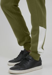 Olivgrüne Jogginghose mit weißen Akzenten, engen Bündchen, getragen mit schwarzen Ledersneakern mit weißen Sohlen und strukturiertem Obermaterial.