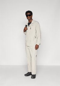 Giacca di un beige chiaro, resistente all'acqua, con colletto alto e tasche con zip, abbinata a pantaloni coordinati e scarpe nere. Modello con occhiali da sole.