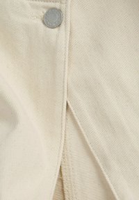 Tela color crema con una sutil textura acanalada, que presenta un botón circular plateado y una solapa de tela con una costura prolija.