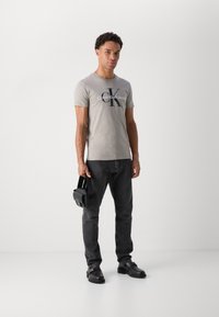 Pelēka kokvilnas t-krekls ar melnu "Calvin Klein Jeans" logo, apvienots ar tumši pelēkiem džinsiem un melnām kurpēm. Tur rokās mazs melns soma.