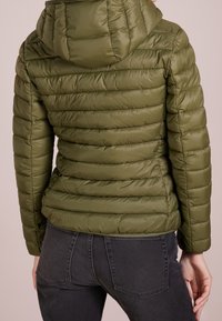 Olijfgroene pufferjas met capuchon, voorzien van een horizontale gewatteerde structuur en elastische manchetten. Gecombineerd met donkere jeans voor contrast.