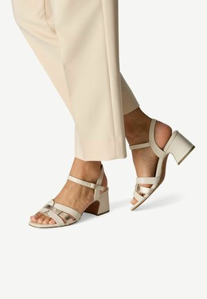 Sandales beiges à brides avec un talon bloc, présentant des brides courbes et un accent métallique. Texture en cuir lisse avec un design minimaliste.