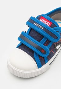 Sneaker in tela blu con punta in gomma bianca, dettaglio di ragno nero e cinturini regolabili in velcro blu. Presenta il marchio "GEOX" e "MARVEL".
