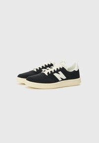 Sorte semskede sneakers har en hvit logo og detaljer, med en teksturert overflate, flate snørebånd og en kremfarget gummisåle.