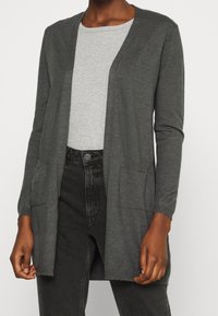 Mörkgrå öppen cardigan med fickor, bärs över en ljusgrå ribbad T-shirt med rund halsringning och svarta högmidjade jeans.