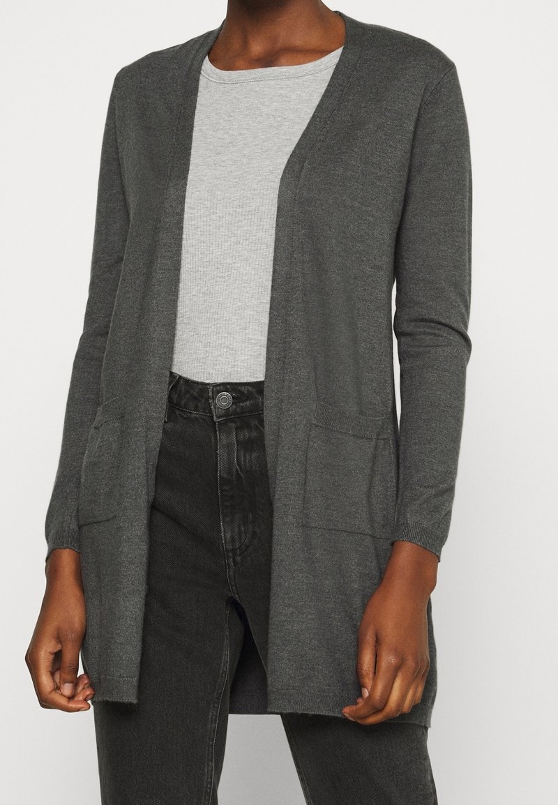 Mörkgrå öppen cardigan med fickor, bärs över en ljusgrå ribbad T-shirt med rund halsringning och svarta högmidjade jeans.