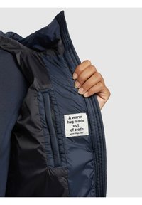 Interior de chaqueta acolchada azul marino con textura suave. Una etiqueta dice: "Un abrazo cálido hecho de tela". Mano sosteniendo el borde de la chaqueta.