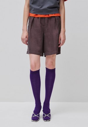 Persona con pantalones cortos marrones, calcetines morados hasta la rodilla y tacones abiertos morados sobre un fondo gris liso.