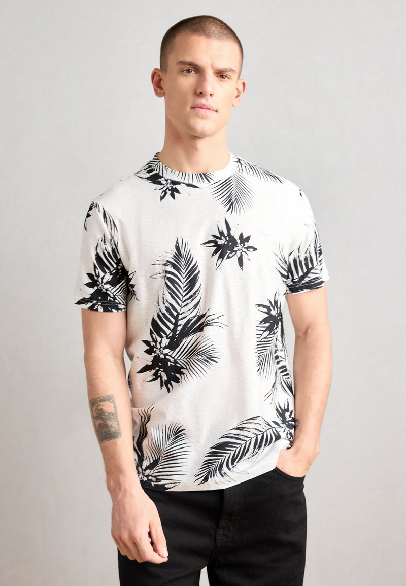 Korte mouwen t-shirt van witte stof met een zwart tropisch bladenpatroon, ronde halslijn en een relaxte pasvorm. Model draagt donkere jeans.