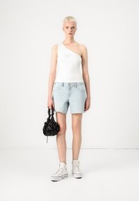 Hvit en-skulder topp, lyseblå denimshorts med frynsete kant, svart mønstrete veske og hvite sneakers med høy såle.