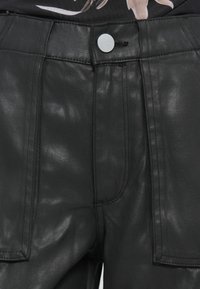 Pantalones negros de material brillante y texturizado con costuras visibles y dos bolsillos delanteros, con un cierre de botón plateado y cremallera.