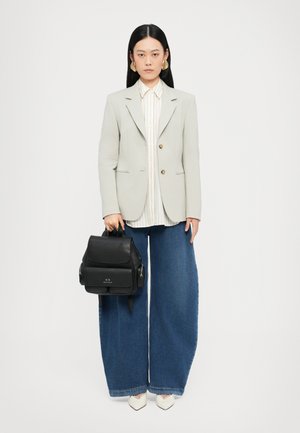 Svart lær ryggsekk med flere lommer, holdt av en person i en lys grå blazer, stripete skjorte og vide blå jeans. Hvite sko.