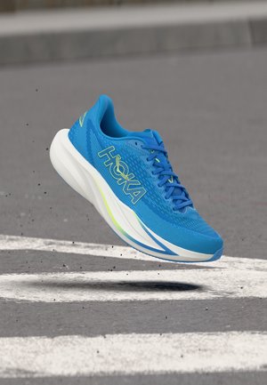 Scarpa da corsa Hoka blu con suola bianca spessa, che fluttua sopra l'asfalto con segnaletica stradale bianca.