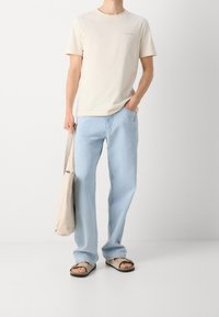 Camiseta de algodón beige claro con un pequeño logo, jeans de mezclilla azul claro, sandalias beige y una bolsa tote beige. Corte relajado y estilo casual.