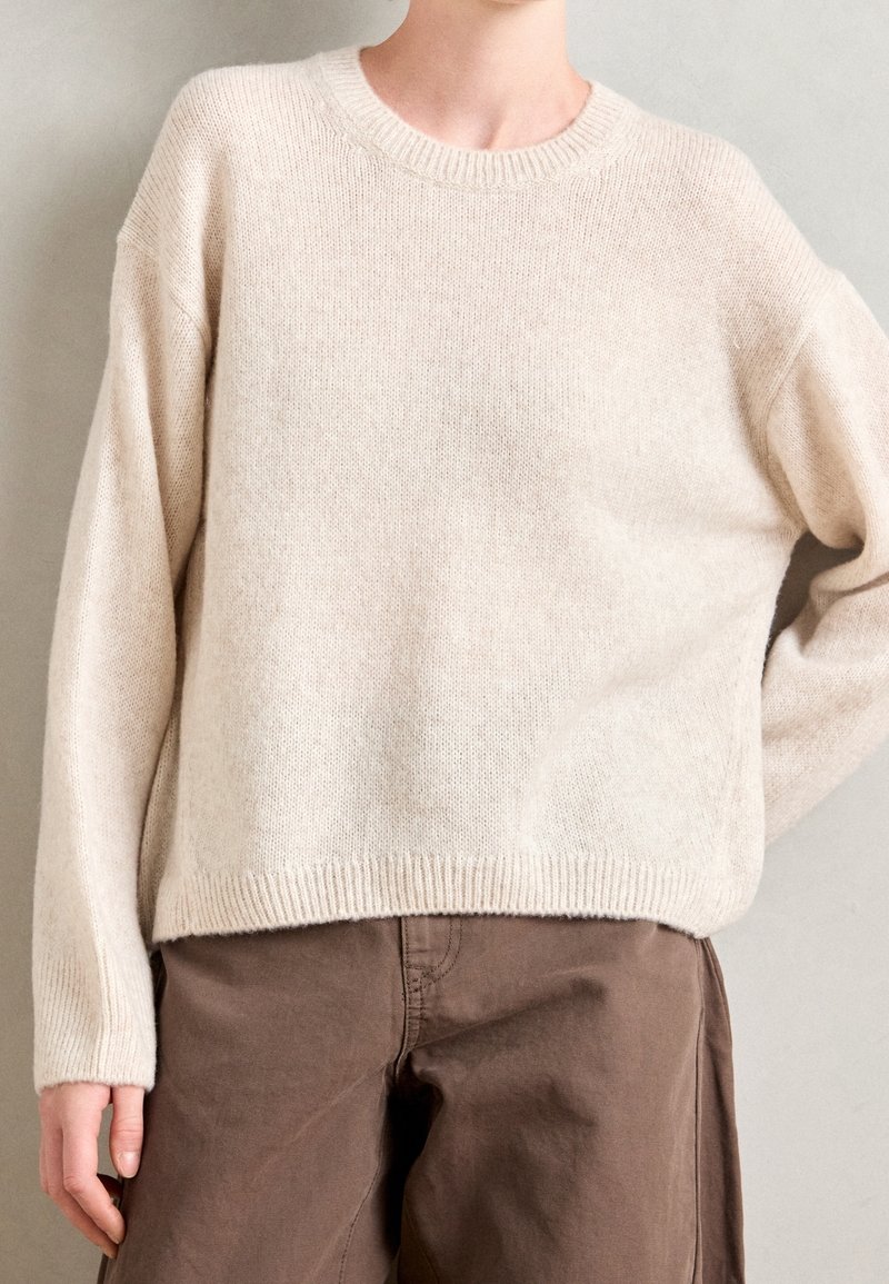 Personne portant un pull en maille ample de couleur crème et un pantalon marron, debout devant un fond uni gris clair.