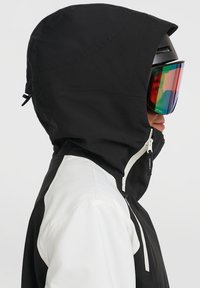 Zwart-witte skijas met een hoge kraag, voorzien van een ritssluitzak en een trekkoord capuchon, gecombineerd met gespiegeld skibril.