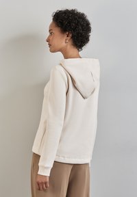 Felpa con cappuccio beige realizzata in tessuto morbido, con una vestibilità comoda e polsini a coste. La vista posteriore non mostra motivi o parti metalliche visibili.