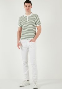 Csíkos zöld és fehér rövid ujjú pólóing gallérral, fehér slim-fit farmernadrággal és fehér sportcipővel párosítva, semleges háttér előtt állva.
