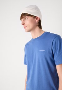 Blue cotton t-shirt with short sleeves, round neckline, and small white "Calvin Klein" logo on the chest. Worn with a white ribbed beanie.