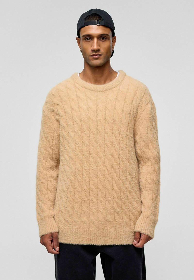 Beige kabelgebreide trui gemaakt van zachte, pluizige stof met een ronde halslijn en lange mouwen, gedragen over donkere broeken.