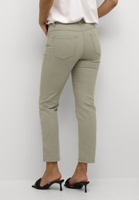 Jeans skinny color caqui con una cintura de media altura, cinco bolsillos y una textura suave, combinados con sandalias de tacón negras.