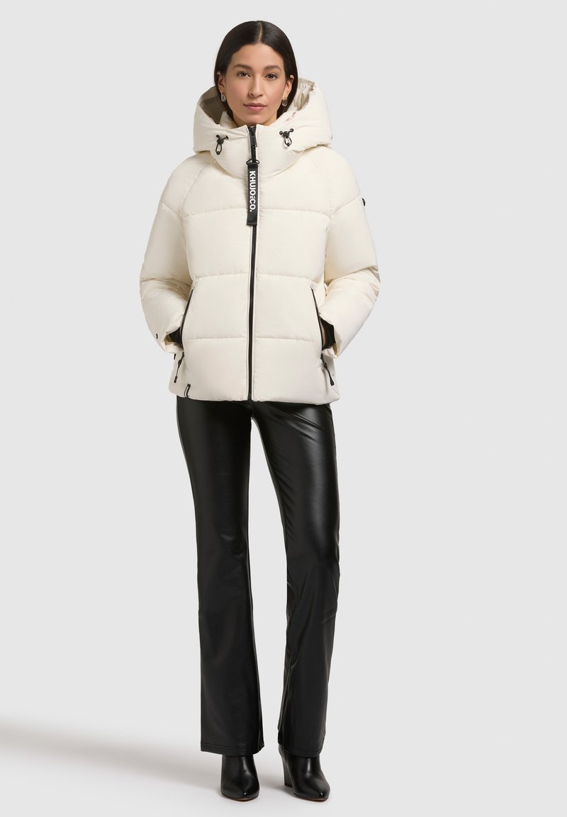 Cremfarbener Puffer-Jacke mit hohem Kragen und verstellbarem Kapuze, ausgestattet mit Reißverschlusstaschen und schwarzen Akzenten, kombiniert mit glänzenden schwarzen Hosen.