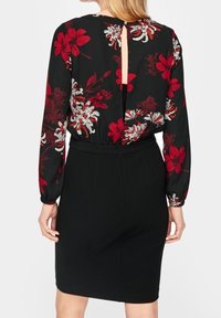 Femme portant une robe noire à manches longues, avec un motif floral rouge et blanc sur le haut, et une petite ouverture en forme de trou à l'arrière.