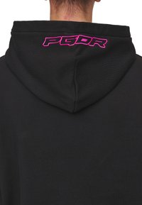 Čierna mikina s kapucňou s výrazným ružovým vyšitým logom „PGDR“ na zadnej strane kapucne. Tkanina vyzerá hladko a má mierne štruktúrovaný povrch.