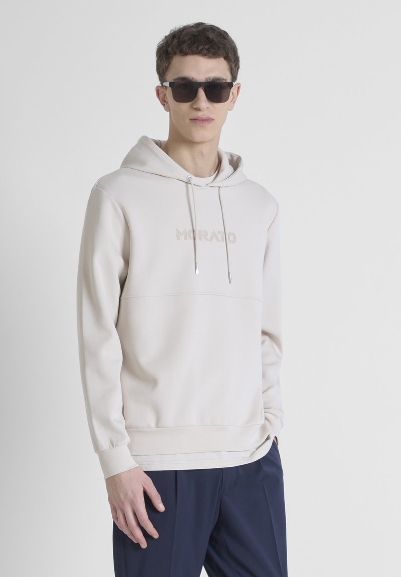 Sudadera beige con cordón, bolsillo canguro en el frente y logotipo en relieve. El modelo lleva gafas de sol negras y pantalones azul marino, contra un fondo blanco.