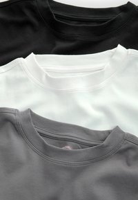 Drie crewneck t-shirts in zwart, wit en grijs, met een gladde stof, ribgebreide kragen en vlakke stiksels langs de naden.