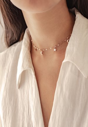 Goudkleurige choker met een gedraaide kettingontwerp, voorzien van twee halve manen en een ster charme, gedragen over een witte, gestructureerde blouse.