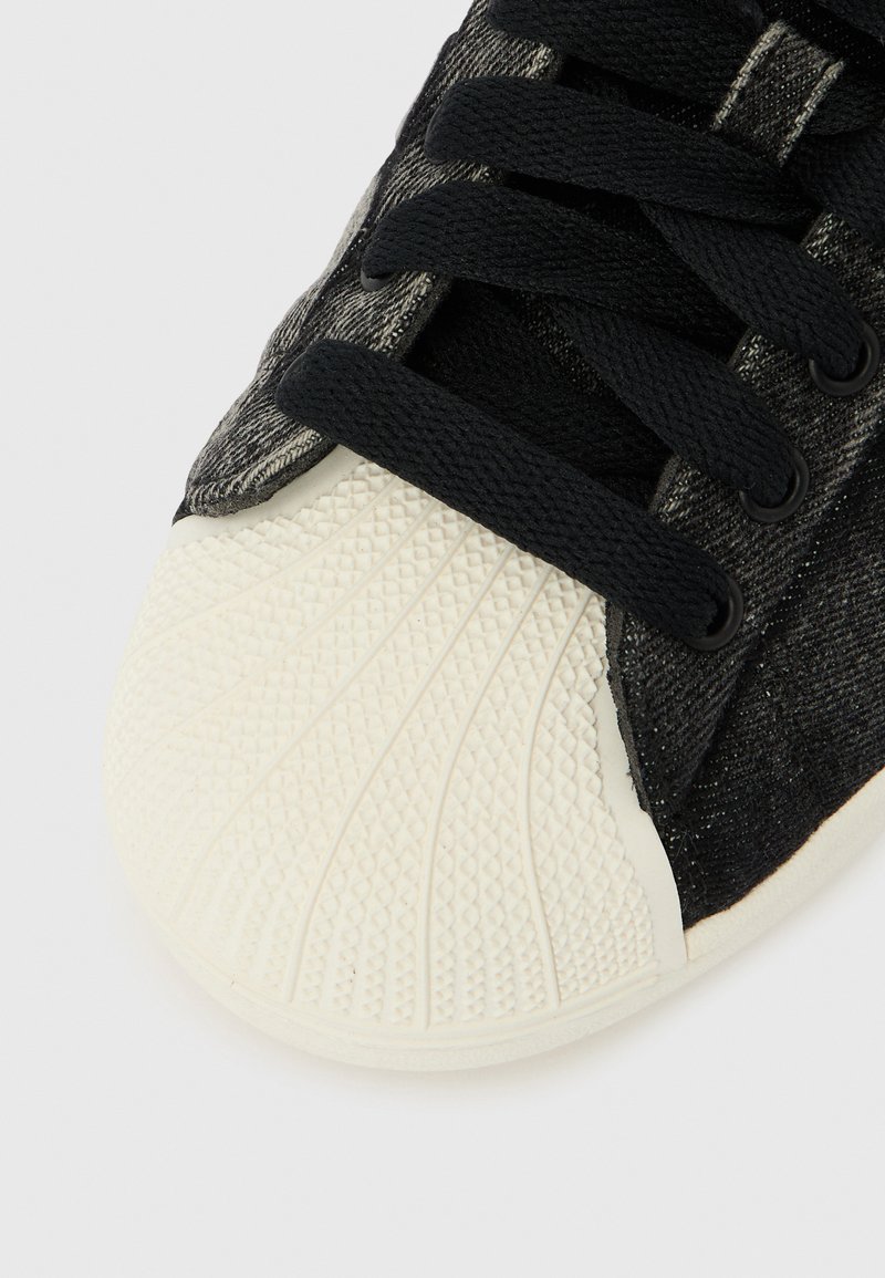 Gros plan d'une basket noire avec un embout en caoutchouc blanc texturé et des lacets noirs sur un fond blanc.