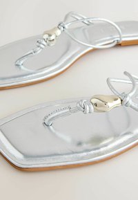 Sandales plates argentées avec des lanières fines et une perle décorative dorée de forme irrégulière sur chaque boucle d'orteil.