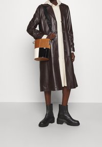Manteau long en cuir marron, robe beige, bottines en cuir noir, et un sac seau à blocs de couleurs avec des sections marron, crème et noir.