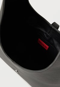 Μαύρο δερμάτινο χέρι-τσάντα ανοιχτό για να δείξει το εσωτερικό από μαύρο ύφασμα και φωτεινή κόκκινη ετικέτα "HUGO" ραμμένη μέσα.