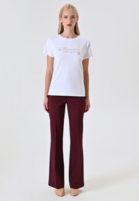 T-shirt bianca in cotone con testo in corsivo dorato e grafica di girasole, abbinata a pantaloni a zampa di elefante borgogna e tacchi beige.