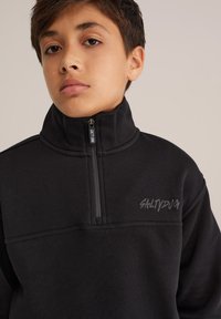 Melnā vilnas jakā ar ceturtdaļas rāvējslēdzēju, augstu apkakli un iespiestu "SALTYDOG" logo uz krūtīm. Iezīmējas ar gludu tekstūru un vieglu dizainu.