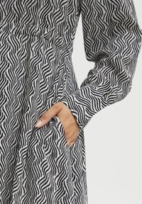 Ilga rankovė suknelė balta su juodais banguotais raštais, su susiraukšlėjusiu liemeniu ir šonine kišenėle, pagaminta iš lengvos medžiagos.