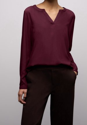 Vrouw draagt een donkerrode V-hals blouse met lange mouwen, ingestopt in een zwarte, hoge taillebroek, met één hand in de zak.