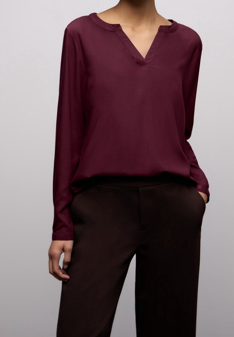 Femme portant un chemisier à manches longues rouge foncé à col en V, rentré dans un pantalon noir taille haute, une main dans la poche.