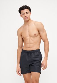 Shorts de bain noirs avec une taille élastique et un cordon de serrage, dotés d'un accent logo sur le côté dans un design ton sur ton subtil. Matière lisse et légère.