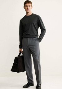 Camicia grigia a maniche lunghe e pantaloni grigi su misura, indossati con scarpe nere. Il soggetto tiene una borsa nera in suede, in piedi contro uno sfondo neutro.