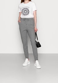 Grå træningsbukser med justerbar snor i taljen, parret med en hvid t-shirt med et sort mandala-print. Hvide sneakers og en lille sort taske.