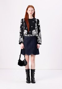 Schwarzes Blümchen-Oberteil mit langen Ärmeln, kombiniert mit einem Jeansrock mit gewelltem Saum. Die Accessoires umfassen eine schwarze Handtasche und knielange schwarze Stiefel.
