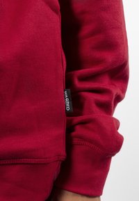 Sudadera roja hecha de una tela suave, con puños acanalados y etiqueta en la costura lateral. La textura parece lisa con un aspecto ligeramente desgastado.