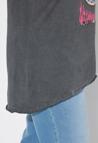 Mørkegrå t-shirt med en let buet kant og synlige rygslids på siderne, prydet med et farverigt grafisk design på fronten. Blå jeans synlige nedenunder.