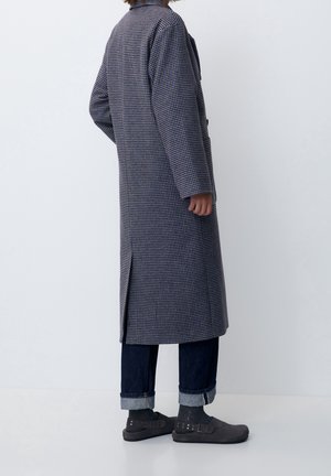 Person iført en lang ternet frakke, opsmøgede mørke jeans og sorte spændesko, stående mod en ensfarvet hvid baggrund.