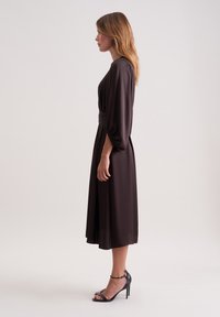 Dunkelbraunes Kleid mit tailliertem Schnitt, langen Ärmeln und fließendem Rock. Kombiniert mit schwarzen High-Heels. Glattes, weiches Material.